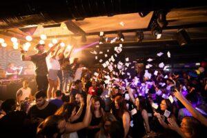 people on stage at a club on campus throwing paper into the crowd of dancing college students